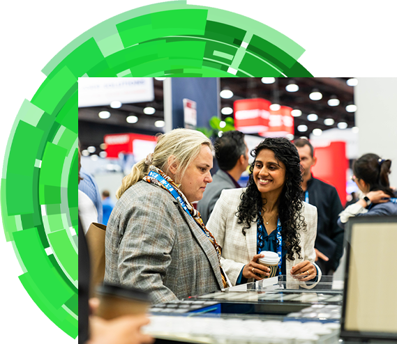 Two women engaged in conversation at a bustling trade show, surrounded by various booths and attendees